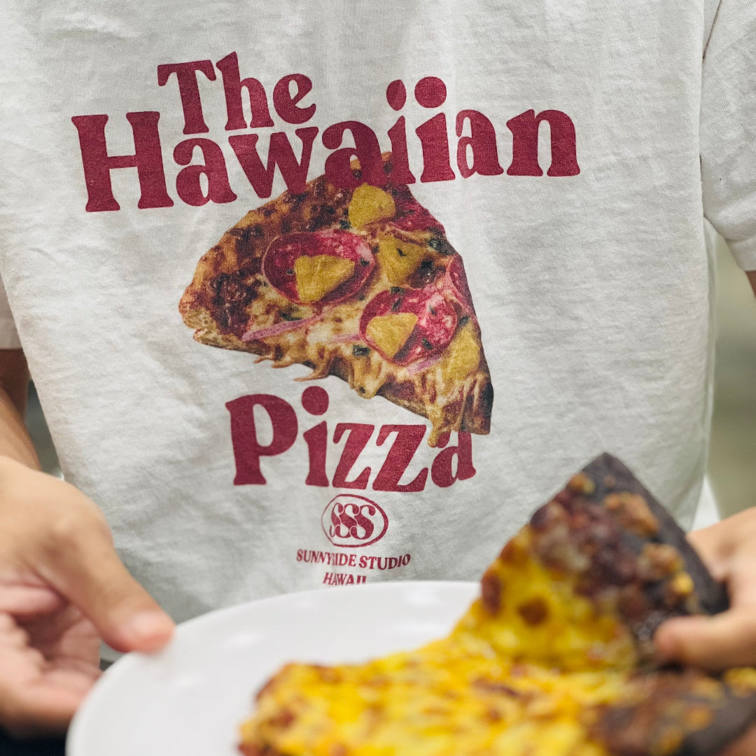 Person wearing a 'The Hawaiian Pizza' t-shirt holding a slice of pizza on a plate.