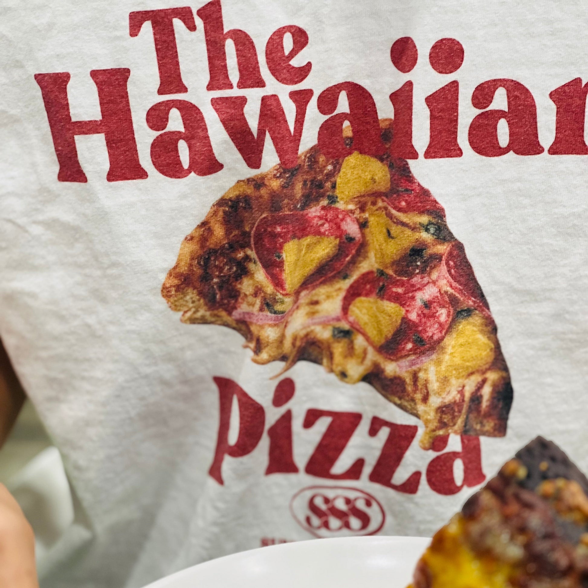 Person wearing a 'The Hawaiian Pizza' t-shirt holding a slice of pizza.