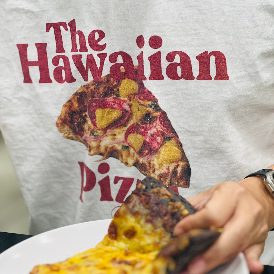 Person holding a slice of pizza with 'The Hawaiian Pizza' t-shirt in the background
