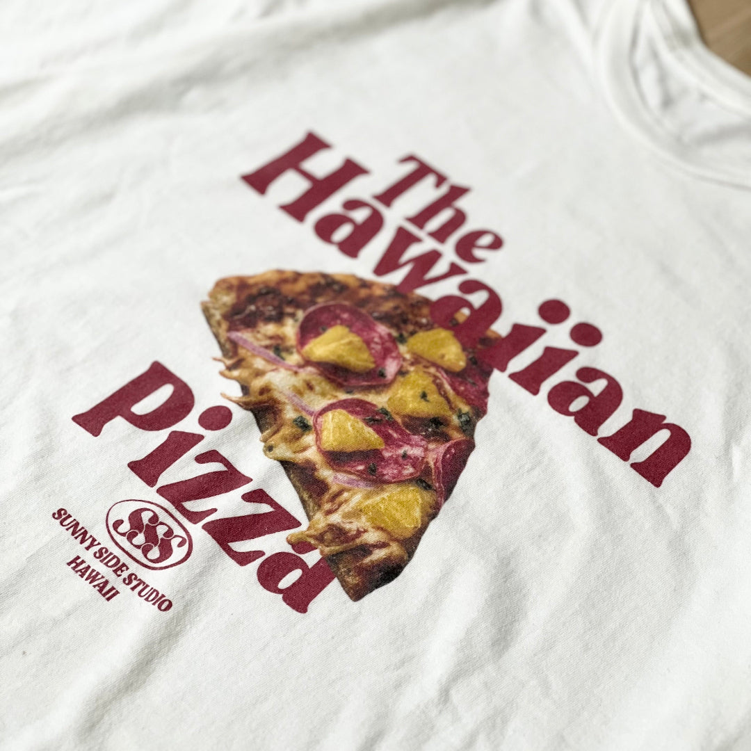 White t-shirt with a pizza slice graphic and text on a wooden surface