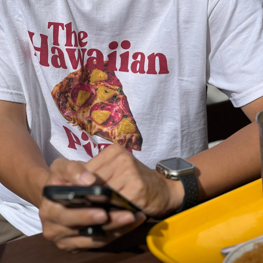 Person wearing a white t-shirt with 'The Hawaiian Pizza' design, holding a phone.